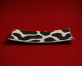 A long, curved ceramic serving platter with an organic black and off-white cow-print pattern, set against a solid red background.