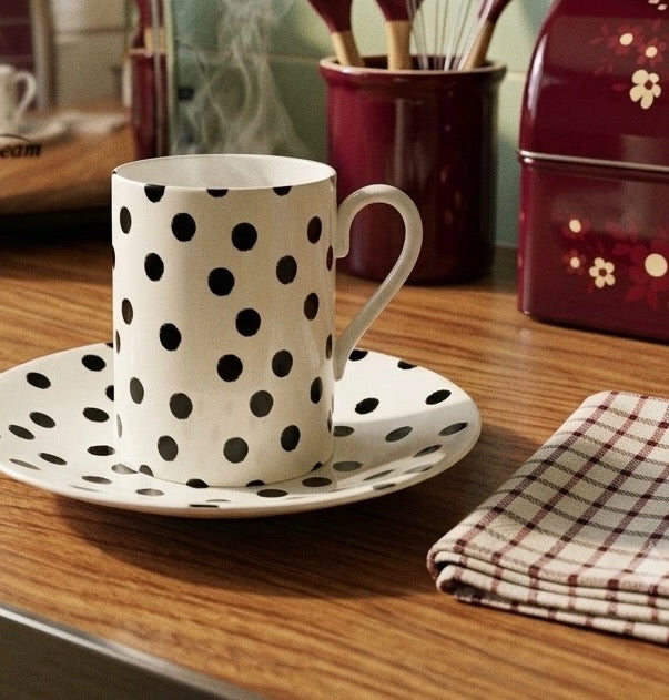 A white ceramic mug with black polka dots sits on a matching saucer on a wooden table, with a folded plaid napkin nearby and kitchen items in the background.