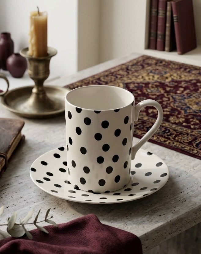 A white ceramic mug with black polka dots sits on a matching saucer on a stone tabletop, styled with a candle, books, and a patterned runner in the background.