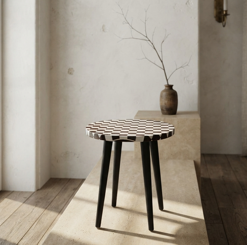 A small round side table with a black and white checkerboard patterned top and black tapered legs, placed in a sunlit minimalist interior with a vase and branch in the background.