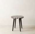A small round stool with a black and white checkerboard patterned top and four dark tapered legs, photographed in a minimal studio setting with a light beige wall and floor.
