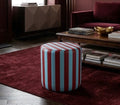 A cylindrical upholstered ottoman with vertical red and light blue stripes, placed on a dark red rug in a styled living room with a sofa, coffee table, and cabinet in the background.