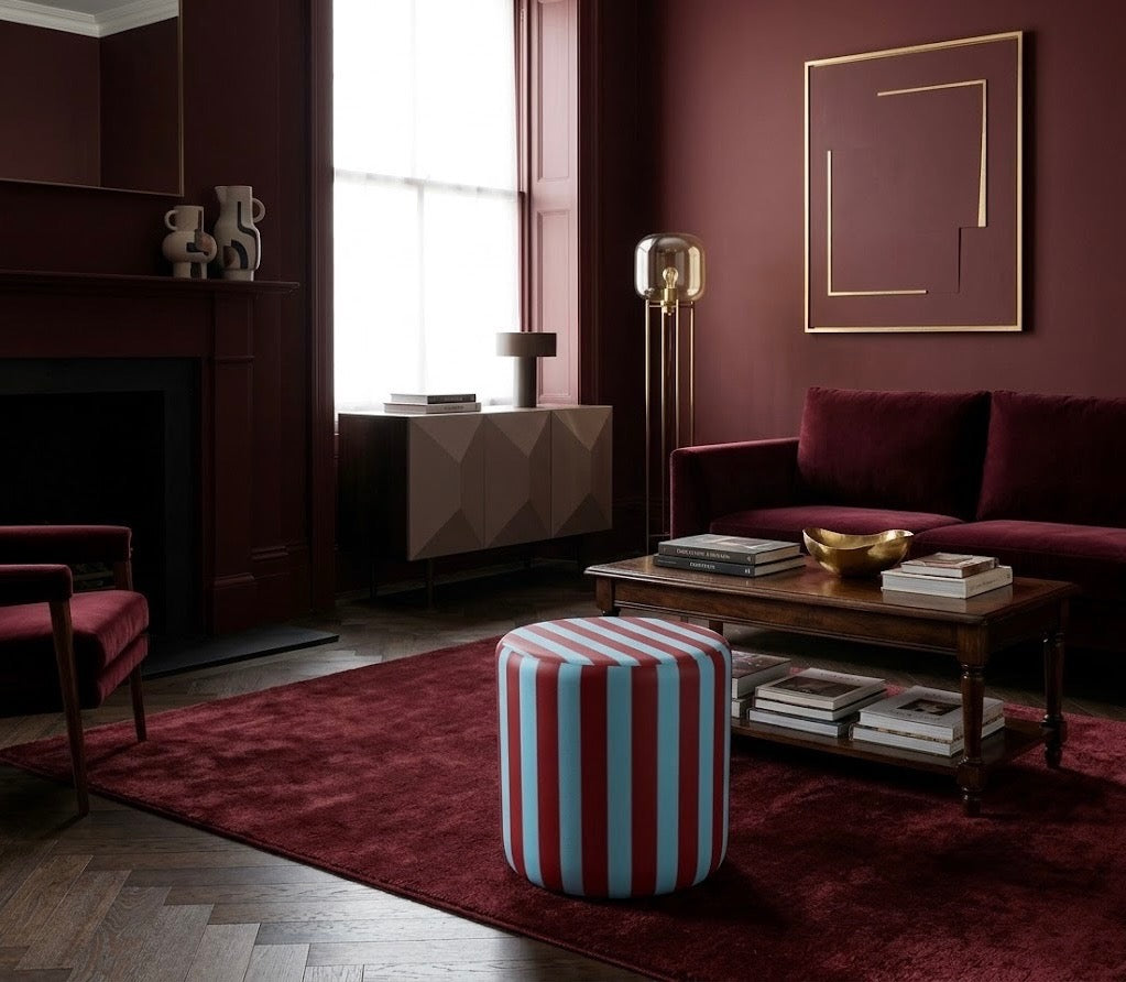A styled living room with deep burgundy walls and furniture, featuring a striped cylindrical ottoman in the foreground, a wooden coffee table, a sofa, and a large framed abstract wall art piece.