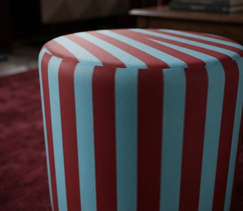 A cylindrical upholstered ottoman or stool with bold vertical red and light blue stripes, shown in a real indoor setting with a dark red rug and furniture blurred in the background.