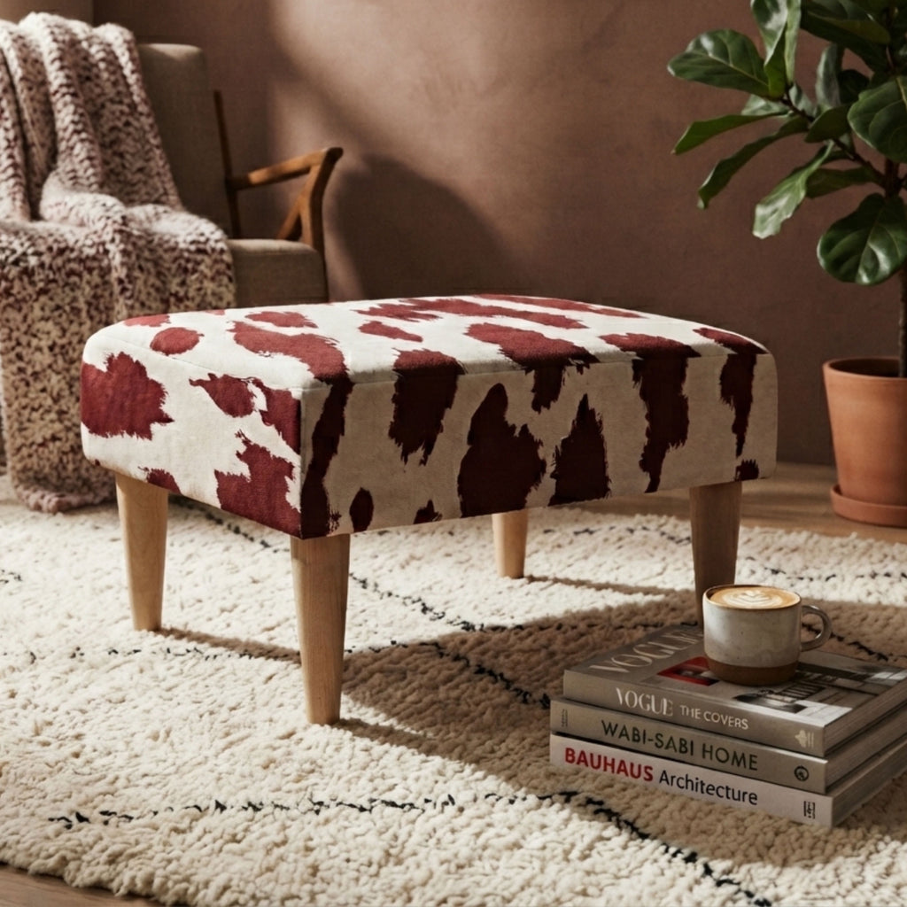 A square upholstered ottoman with a cow-print pattern in dark red and cream, supported by four tapered wooden legs, styled in a cozy living room setting.