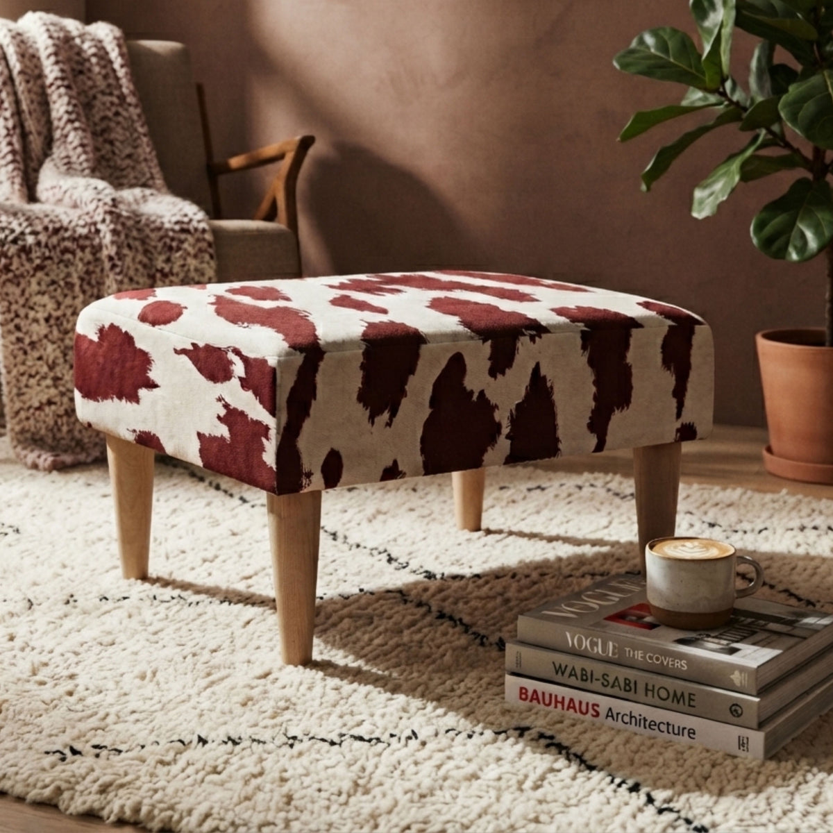 A square upholstered ottoman with a cow-print pattern in dark red and cream, supported by four tapered wooden legs, styled in a cozy living room setting.