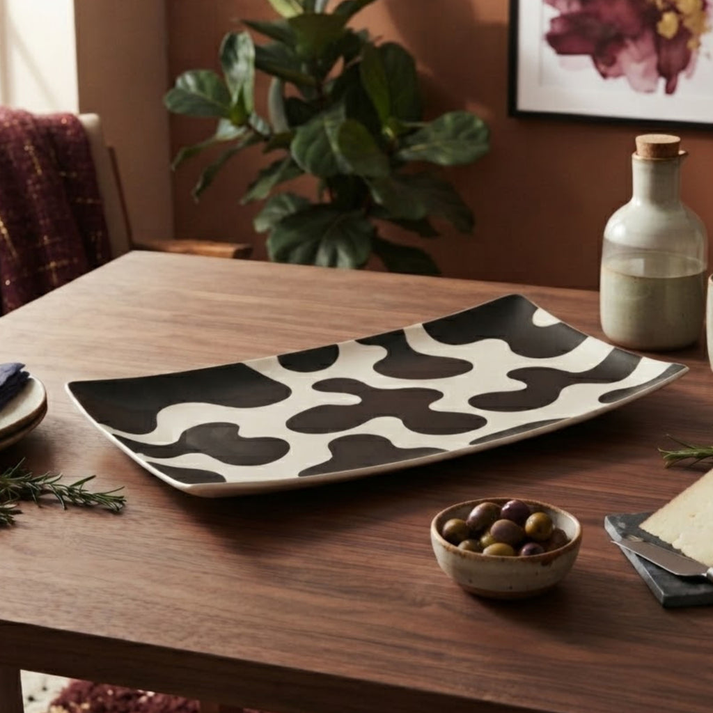 A rectangular ceramic serving platter with a black and white organic cow-print pattern, displayed on a wooden dining table with food props and a plant in the background.