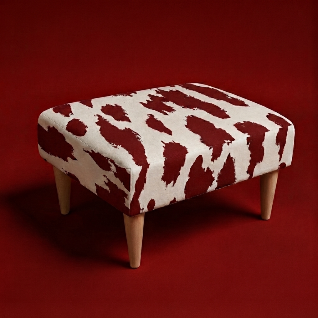 A square upholstered ottoman with a bold red and off-white cow-print pattern and four tapered light wood legs, set against a solid dark red background.