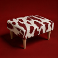 A square upholstered ottoman with a bold red and off-white cow-print pattern and four tapered light wood legs, set against a solid dark red background.