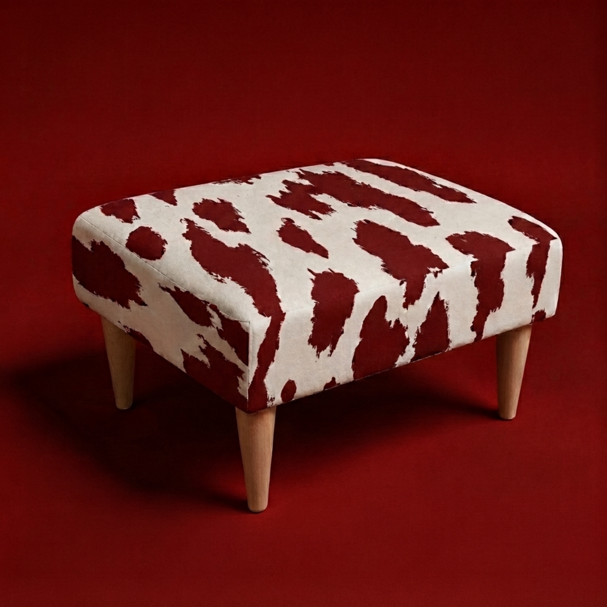 A square upholstered ottoman with a bold red and off-white cow-print pattern and four tapered light wood legs, set against a solid dark red background.