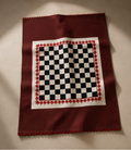 A square burgundy scarf laid flat on a neutral beige background, featuring a central white square with a black-and-white checkerboard pattern and a red decorative border.