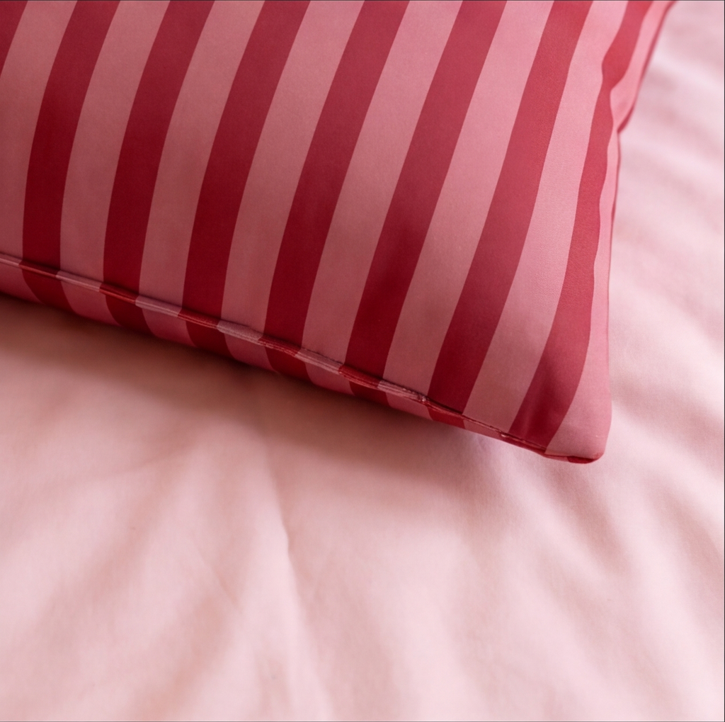 A close-up of a rectangular pillow with vertical red and pink stripes, resting on light pink bedding.