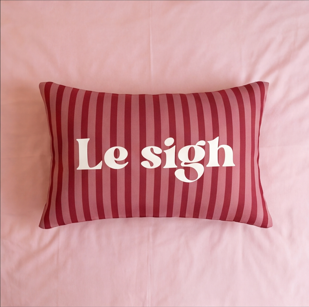 A rectangular throw pillow with red and pink vertical stripes and the phrase 'Le sigh' printed in white serif letters, resting on a pink bed.