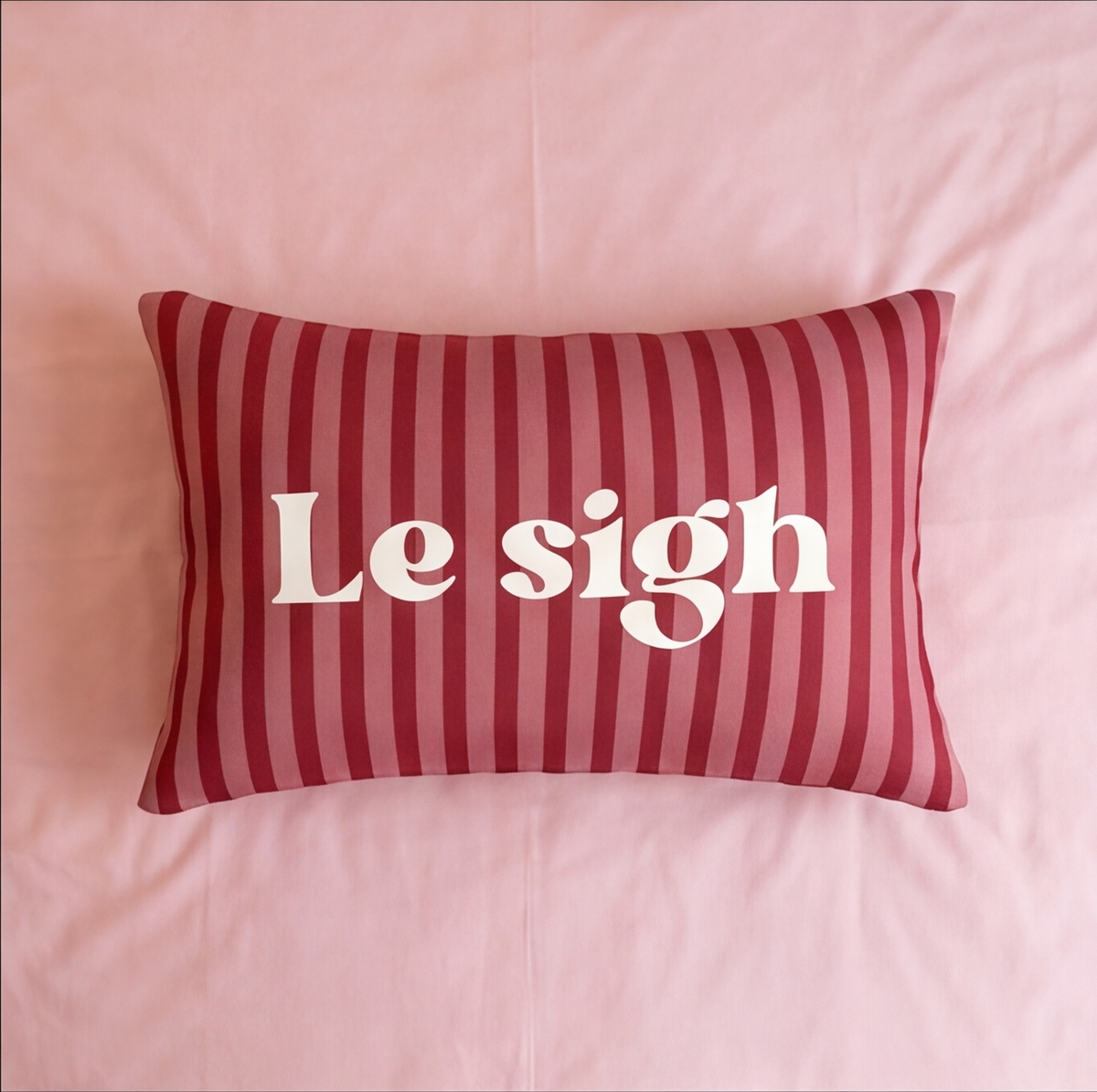 A rectangular throw pillow with red and pink vertical stripes and the phrase 'Le sigh' printed in white serif letters, resting on a pink bed.