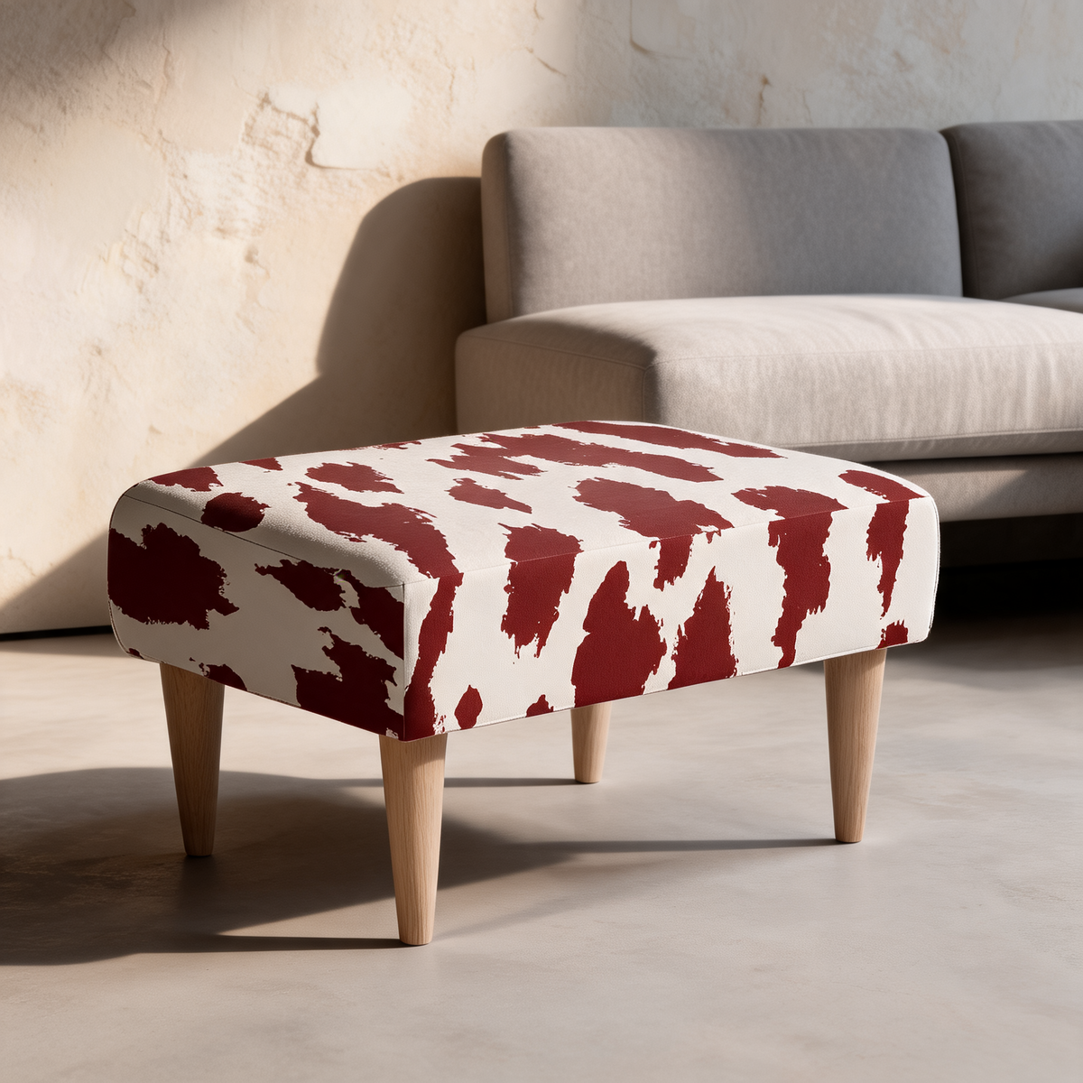 A square upholstered ottoman with a cream base and bold dark red abstract pattern, supported by four tapered light wood legs, set in a modern living room next to a gray sofa.
