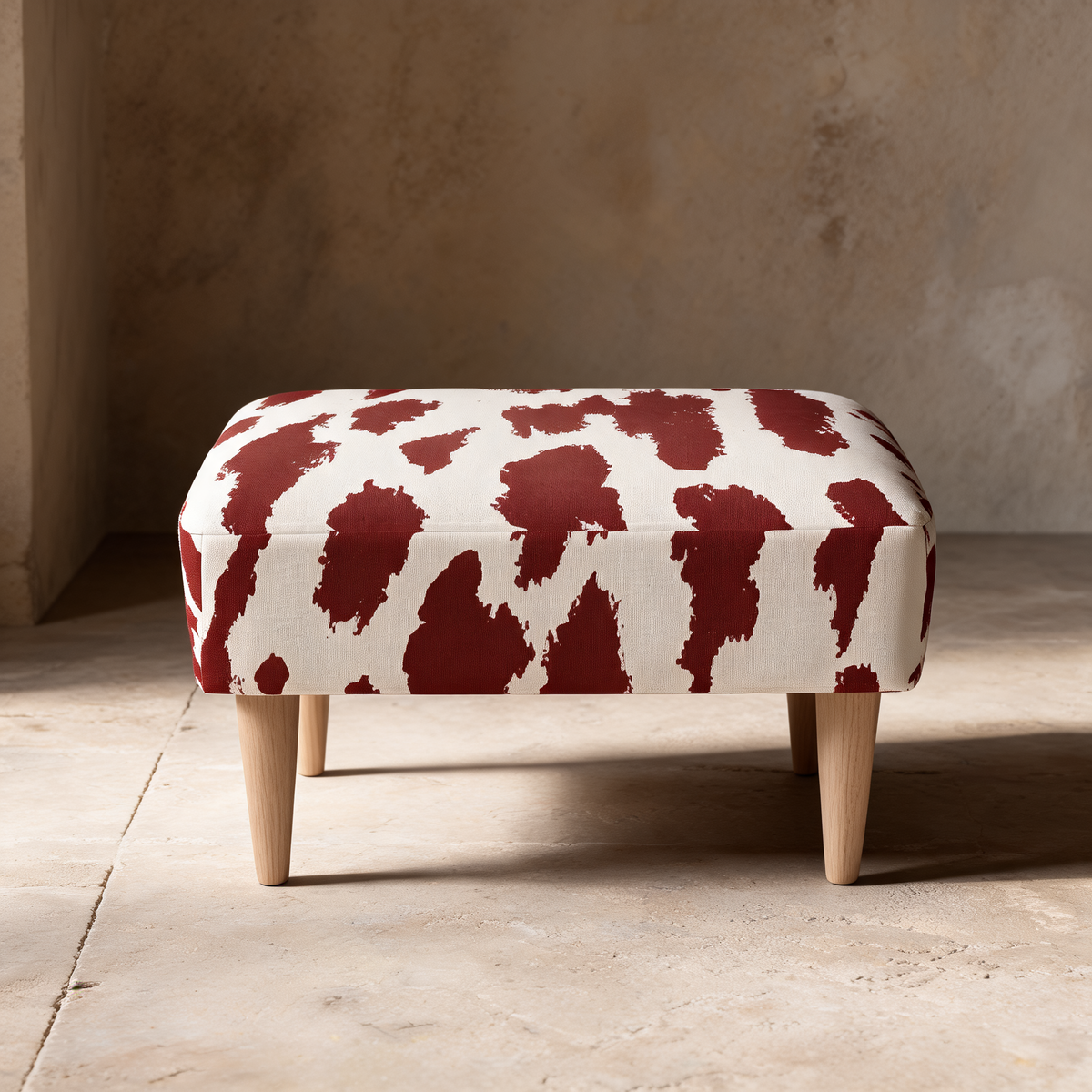 A low rectangular upholstered ottoman with a red and off-white cow-print pattern and light natural wood legs, set against a rustic beige stone wall and floor.