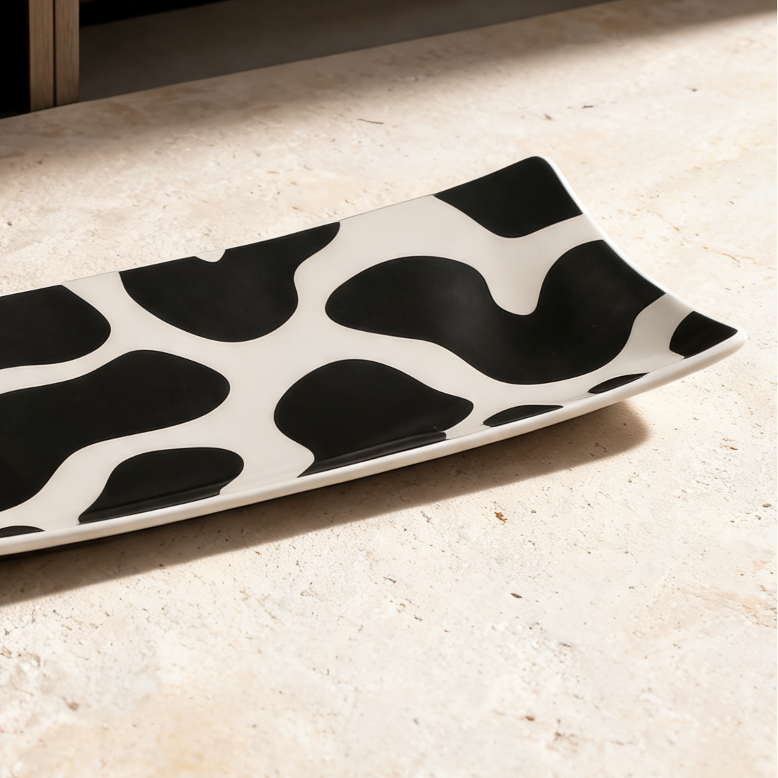 A rectangular ceramic serving platter with a bold black and white organic cow-print pattern, displayed on a textured beige stone surface with a wooden shelf in the background.