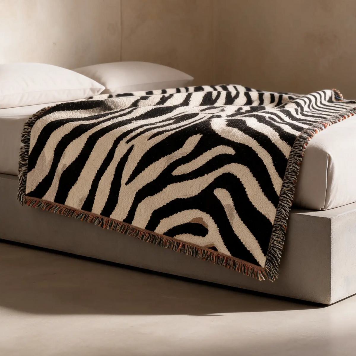 A zebra-print throw blanket with fringe is draped over a modern low-profile bed with white bedding in a minimalist room with textured beige walls and floor.